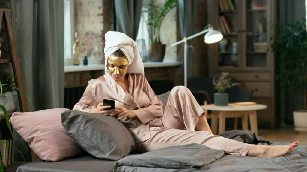A woman in silk pajamas and a gold mask is lying on a bed looking at a phone.