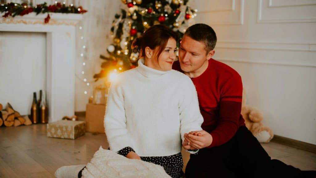 Couple sitting on the floor, holding hands, in front of a Christmas tree and fireplace.