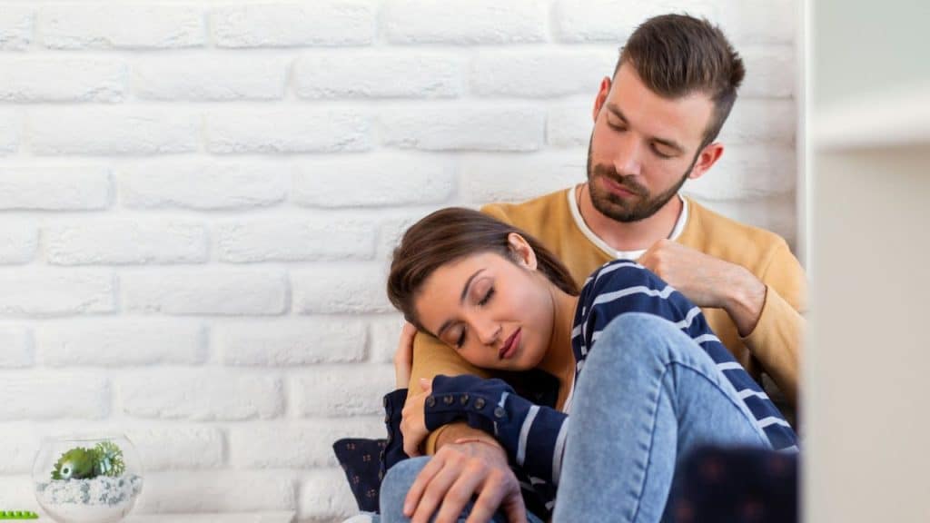A young woman rests her head on a man's chest as he holds her near a white brick wall.