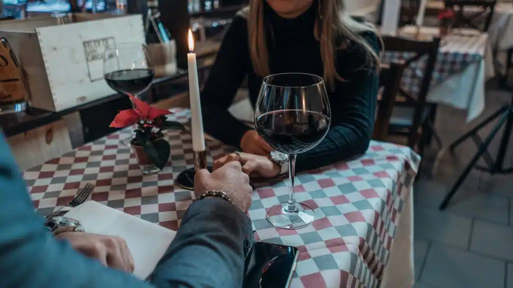A man and a woman are seated at a restaurant table with a lit candle and a glass of red wine, holding hands over the checkered tablecloth.