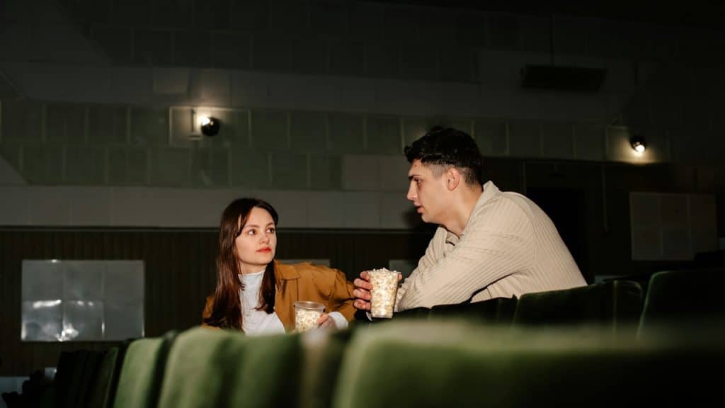 Two people sit in a movie theater, holding popcorn and talking to each other.