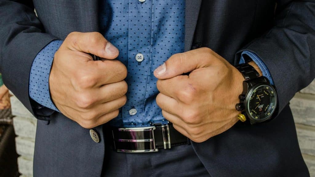 A man in a suit adjusting his jacket, showing his hands, watch, and belt.
