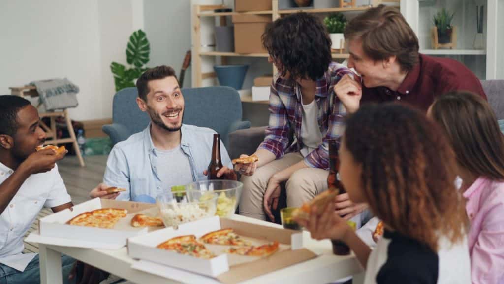 A group of friends sits around a table in a living room, eating pizza and laughing.