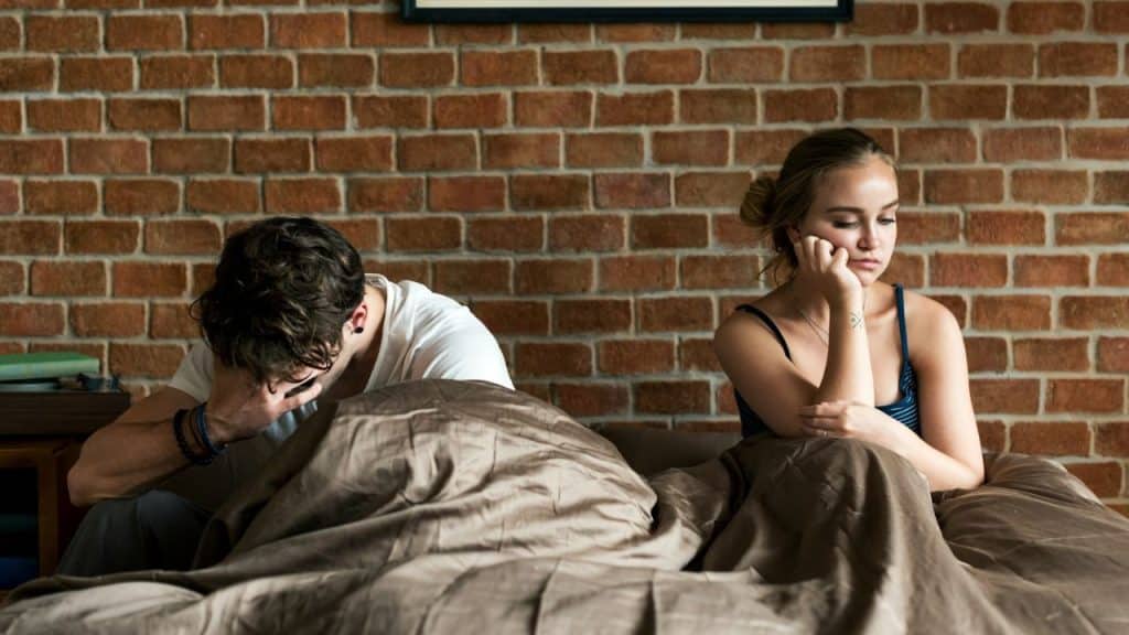 A man is looking down, covering his face while a woman sits thoughtfully on a bed.