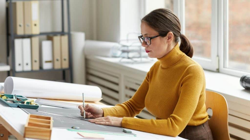 Woman in a yellow turtleneck and glasses sketching at a large desk.