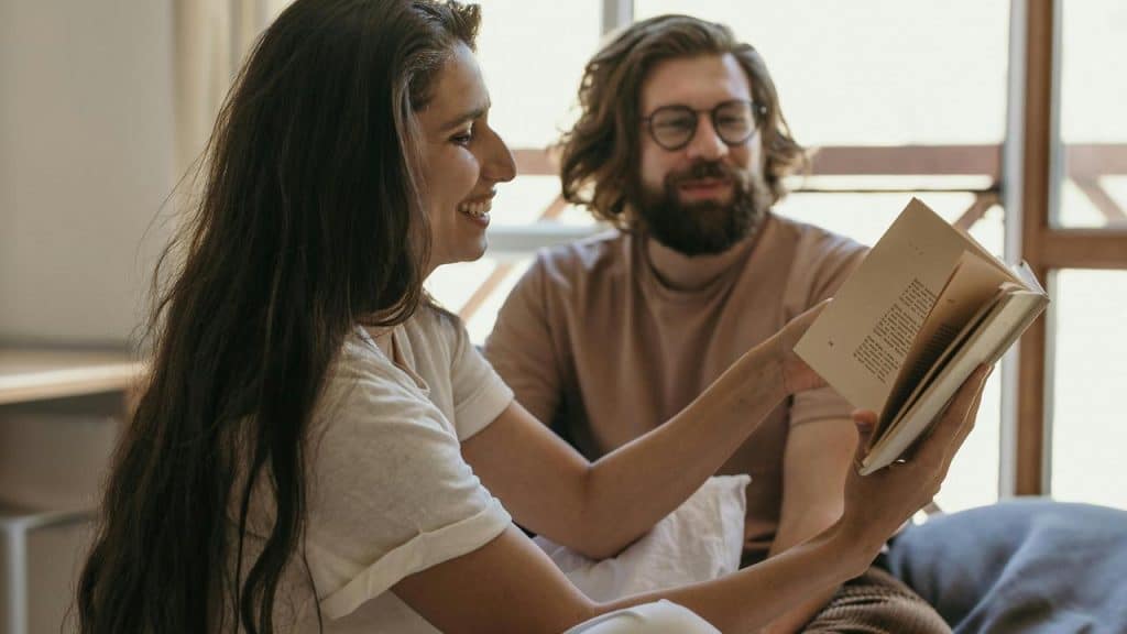 A woman smiling while showing a book to a man sitting beside her.