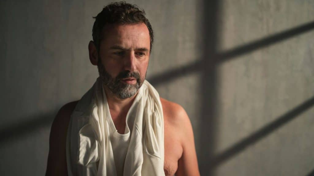 A man in a white undershirt and towel around his neck looks down sadly in soft window light.