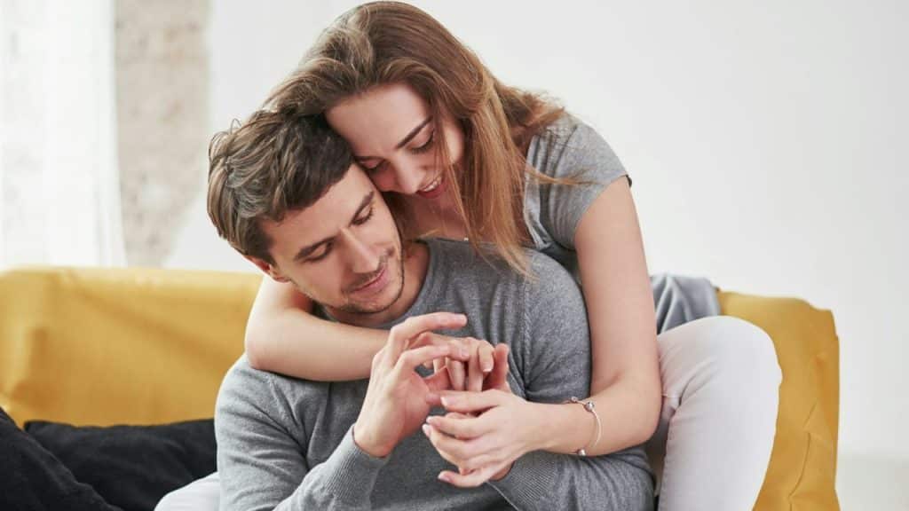 Woman hugging a man from behind, holding his hands, as they sit together.