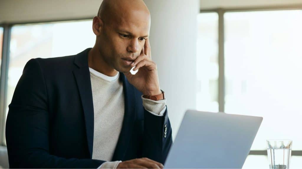 A bald man in a suit jacket concentrates on his laptop, hand resting on his cheek.
