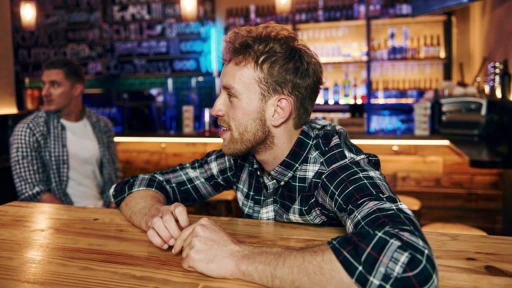 A man in a plaid shirt sits at a bar counter, leaning forward and looking to the side.