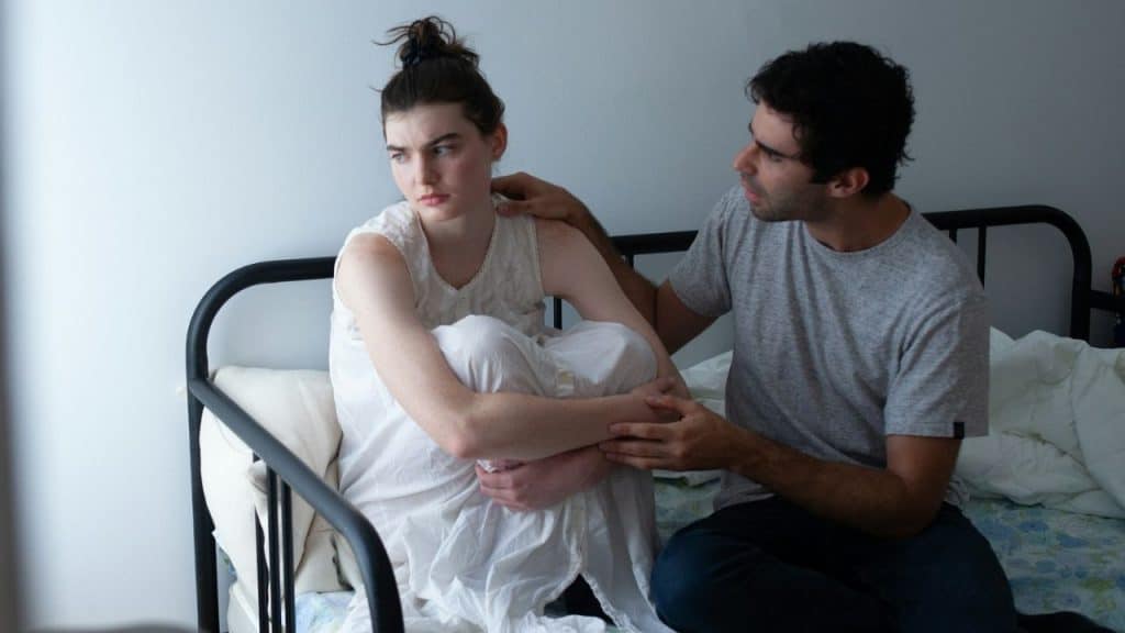 Man gently touches a woman sitting on a bed, who looks distressed and withdrawn.