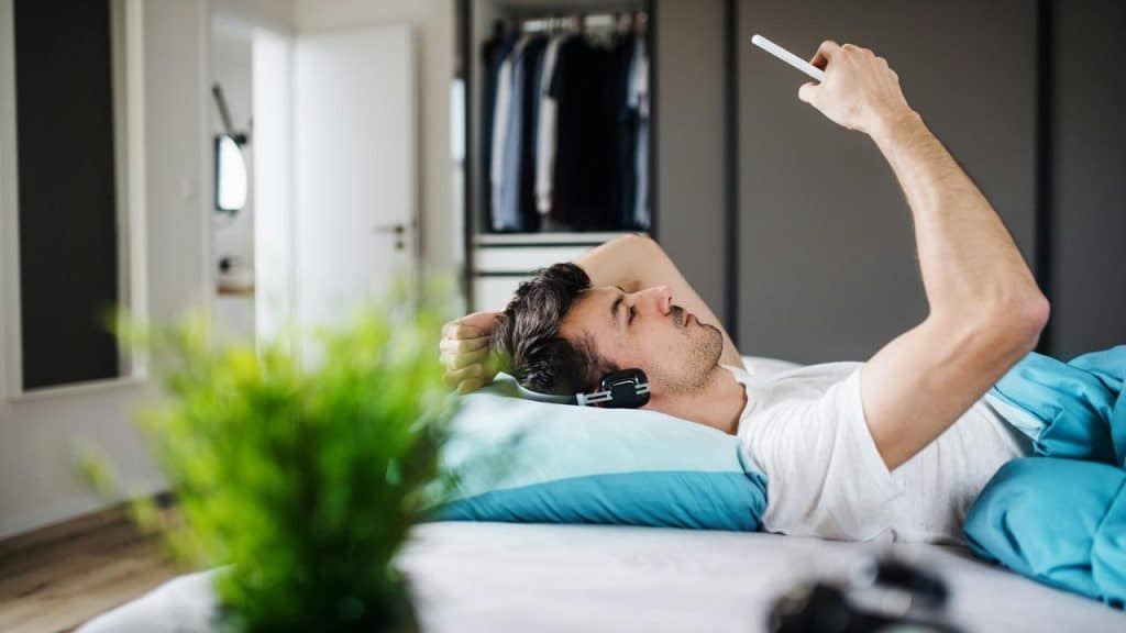 A man lying in bed with headphones on, holding up a smartphone.