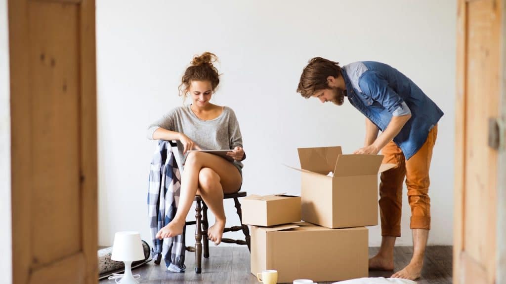 A woman sitting on a chair uses a tablet while a man stands over open cardboard boxes, both barefoot, in a bright room.