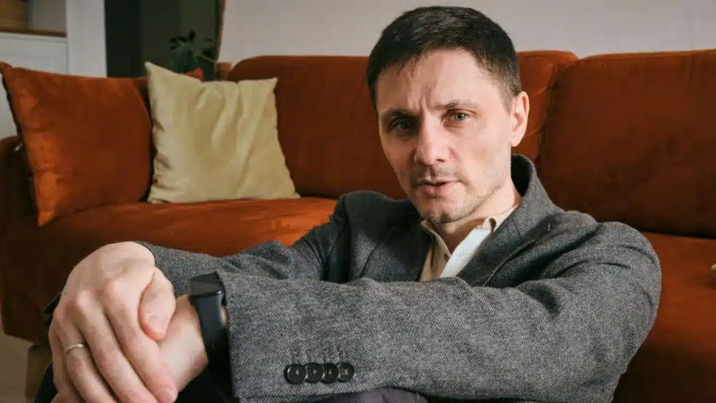 Man in gray jacket and white shirt sitting on floor with an orange couch behind him.