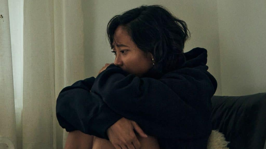 Woman in a black hoodie is sitting, hugging her knees with a sad expression.