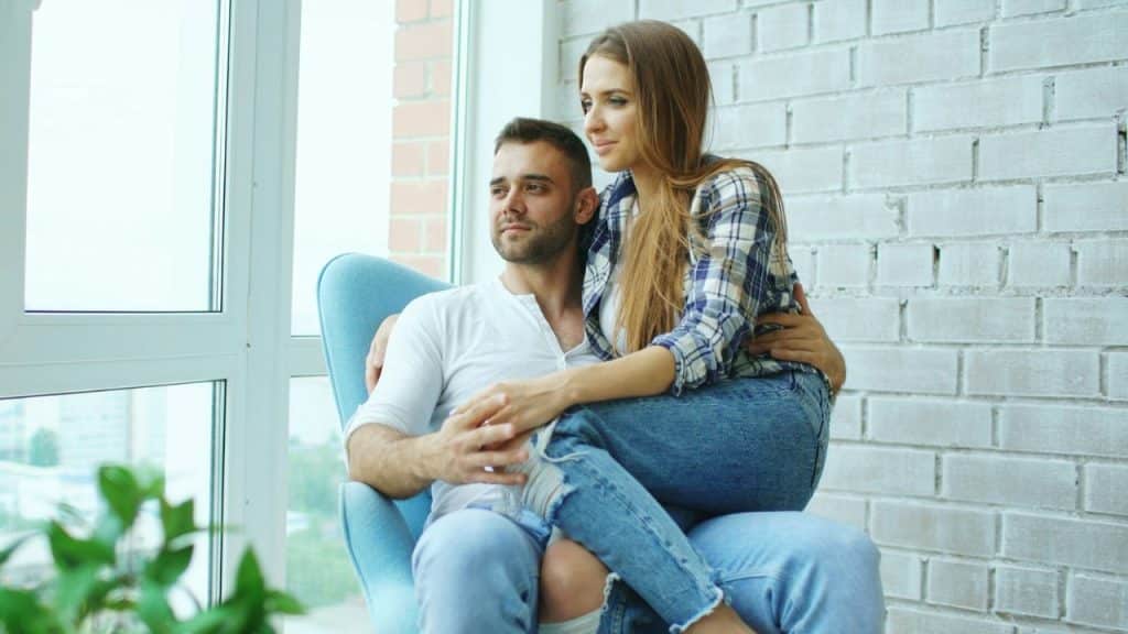 A couple sits by a window, the woman resting on the man's lap.