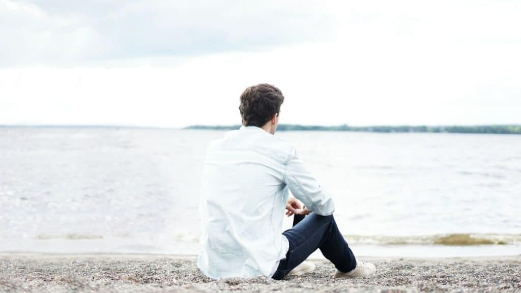 A man is sitting alone on a beach, looking out at a large body of water.