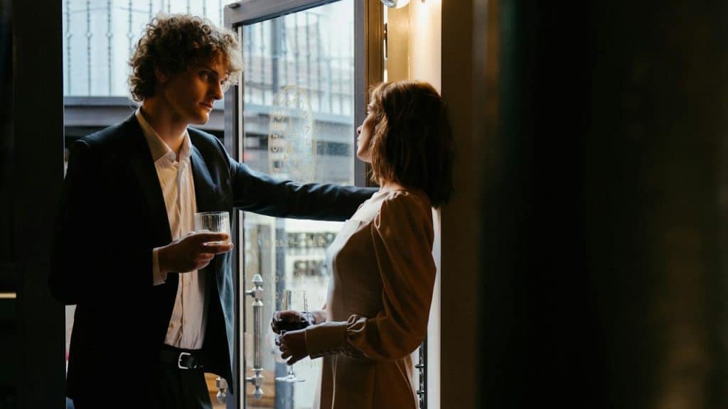 A man in a suit and a woman in a dress talking intimately near a glass door, holding drinks.