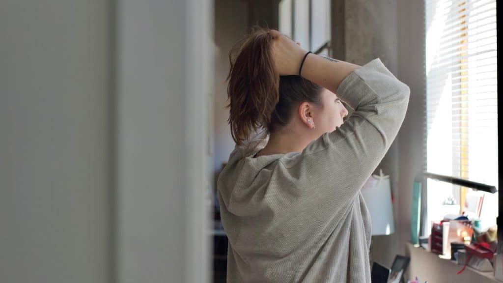 A woman standing by a window tying her hair back.