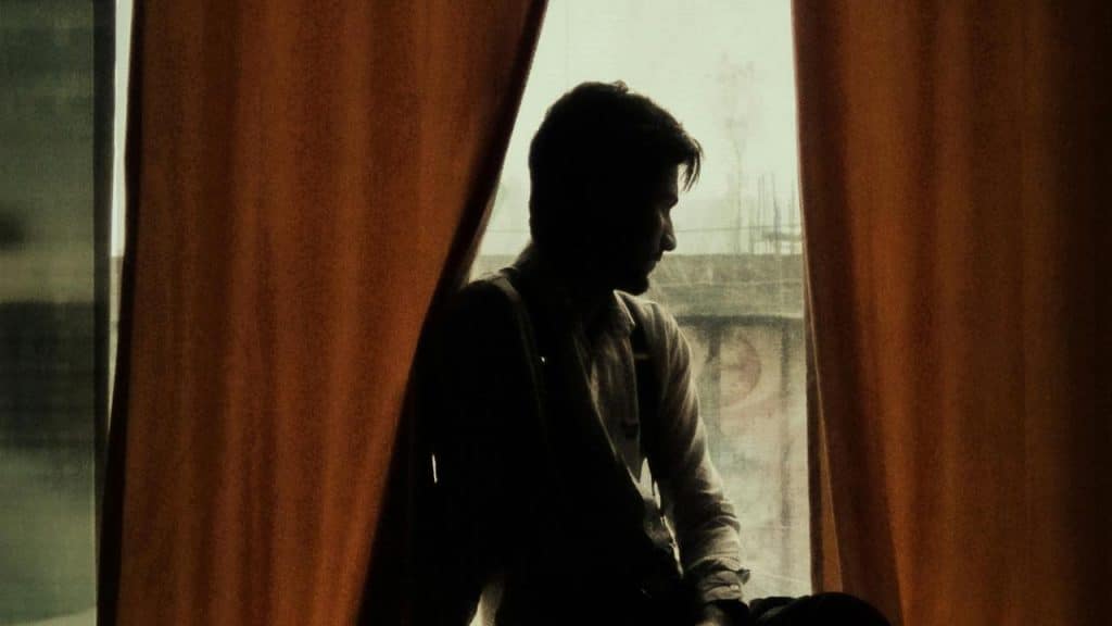 A man sitting by a window framed by curtains, looking outside thoughtfully.