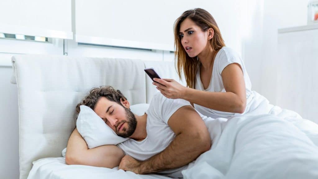 A woman looks at a phone with a shocked expression while sitting next to a sleeping man in bed.