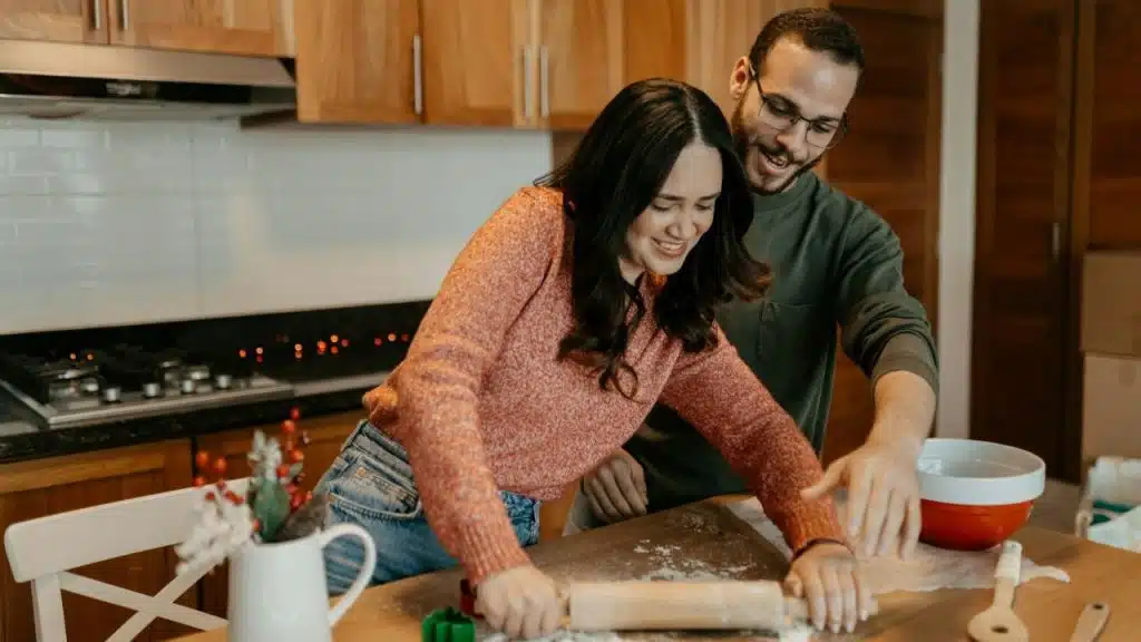 A man and woman are smiling and rolling dough together on a wooden kitchen counter.