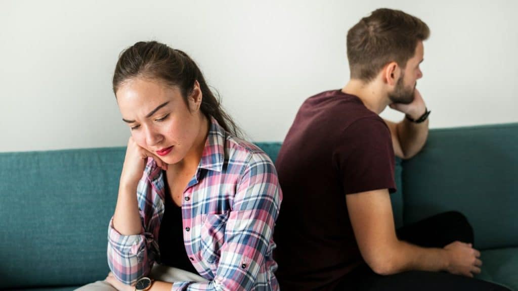 A woman in a plaid shirt sits, looking down sadly, with a man turned away beside her.