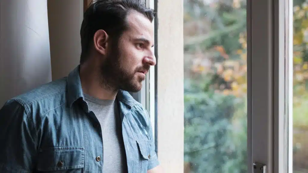 Man with beard and denim shirt looking thoughtfully out a large window.