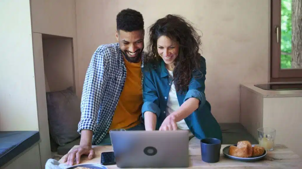 A woman in a blue shirt points at a laptop with a man smiling next to her indoors.