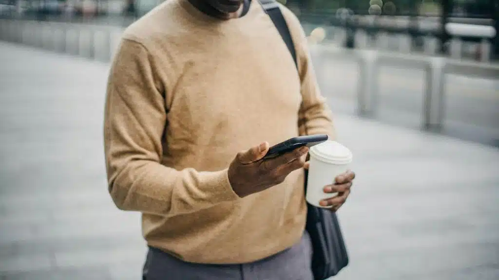A man holding a coffee cup and looking at his phone while standing outdoors.