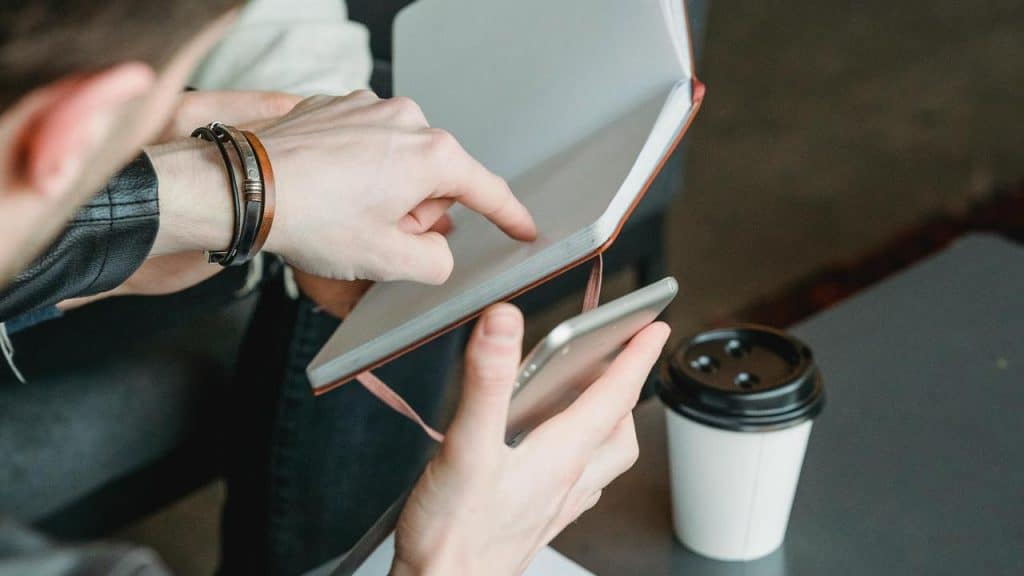A person holding a phone and pointing at an open notebook beside a coffee cup.