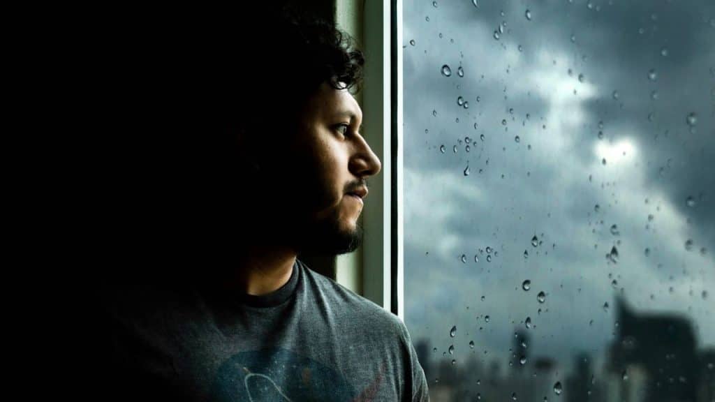 Man in profile looking out a rain-streaked window at a dark, cloudy sky.
