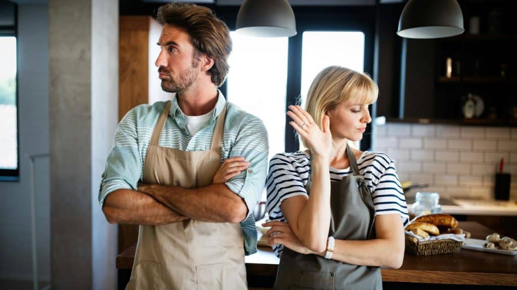 A couple in aprons stand back-to-back in a kitchen, both with arms crossed and looking away.