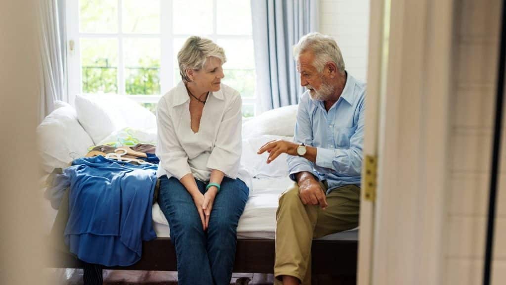 Elderly man sitting on a bed and talking to a woman with clothes beside them.