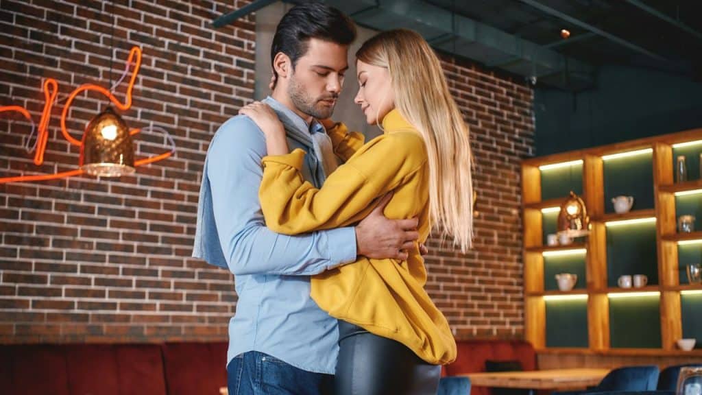 Mature couple embracing, standing close indoors, with a brick wall and shelves behind them.