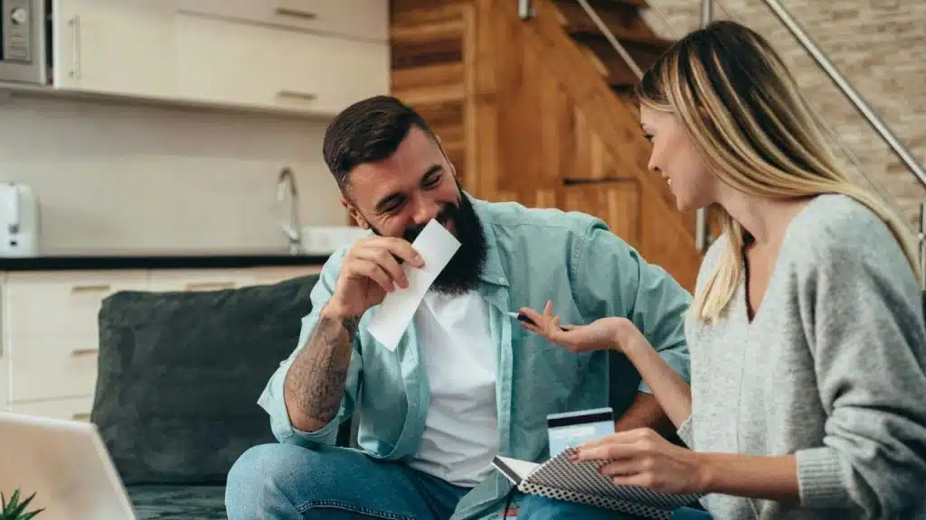 A man laughs, covering his mouth with a slip of paper, while a woman holds a credit card and speaks to him.