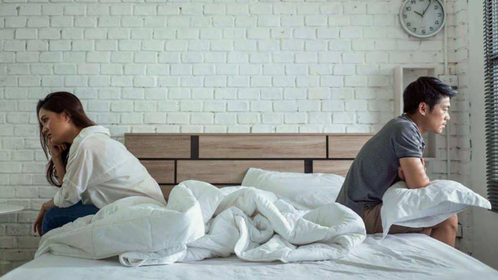 A man and a woman sit back-to-back on a bed, looking distant from each other.