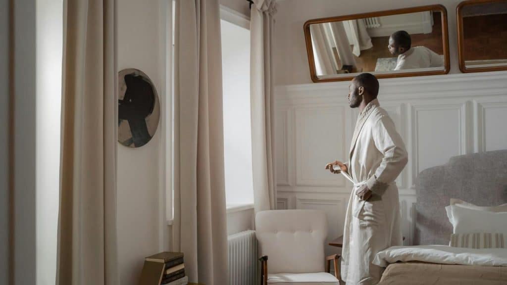 A man in a bathrobe standing by a window looking outside.