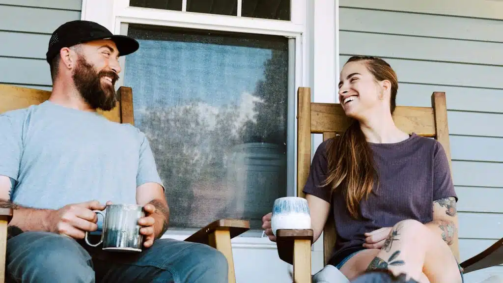 A happy couple sitting on a porch in wooden rocking chairs, smiling and holding mugs.