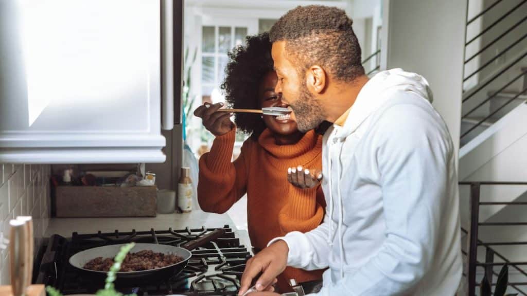 A man in a white hoodie tastes food from a pan offered by a woman with an Afro.