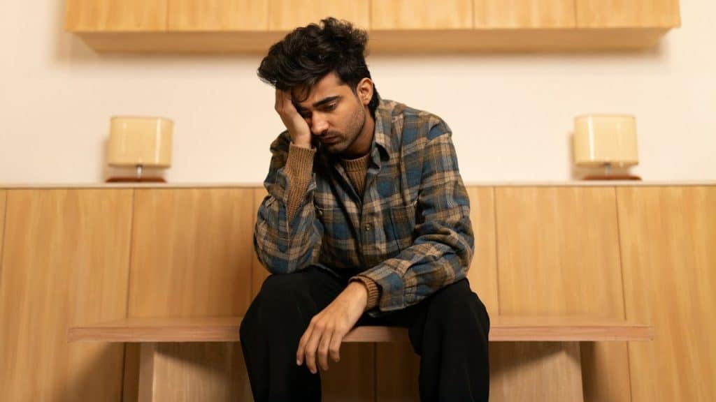 Young man sitting, looking down sadly, with his hand resting on his face.