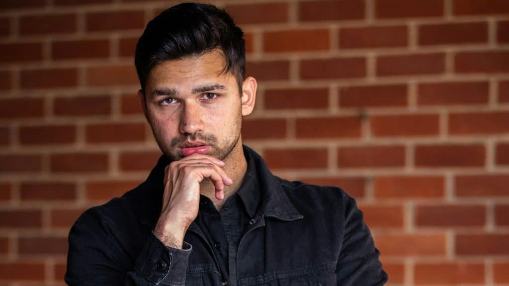 Man in a black shirt jacket against a brick wall, chin resting on his hand.