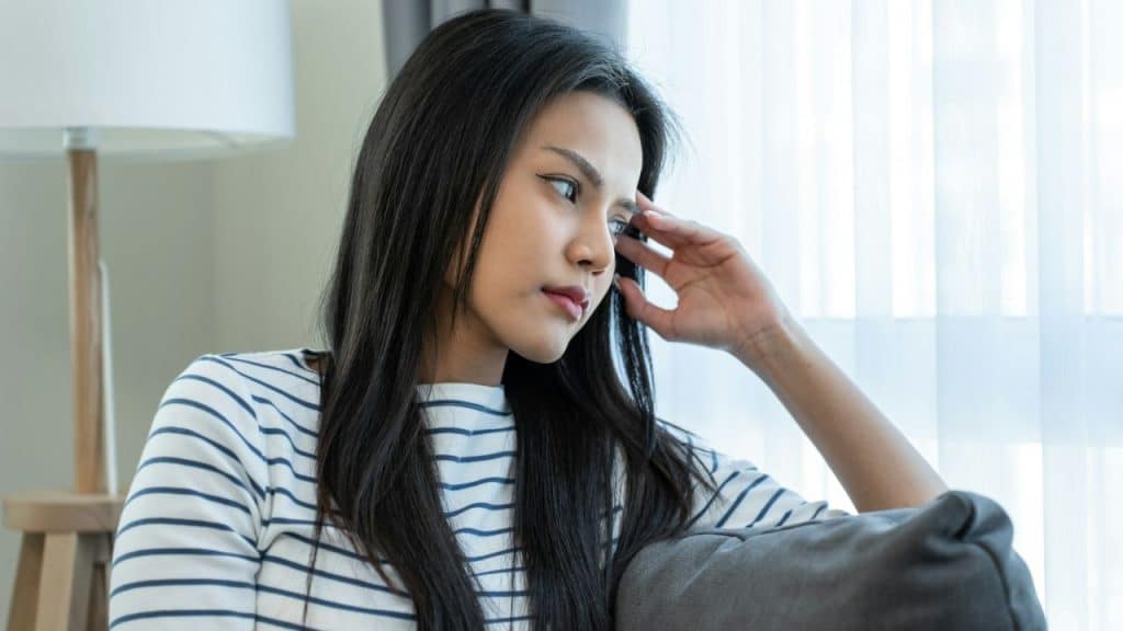 Woman with long black hair and striped shirt sitting on a gray couch looking away.