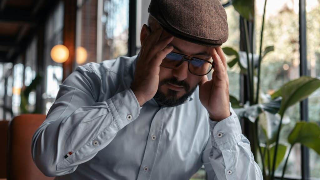 A man wearing glasses and a cap holding his head indoors.