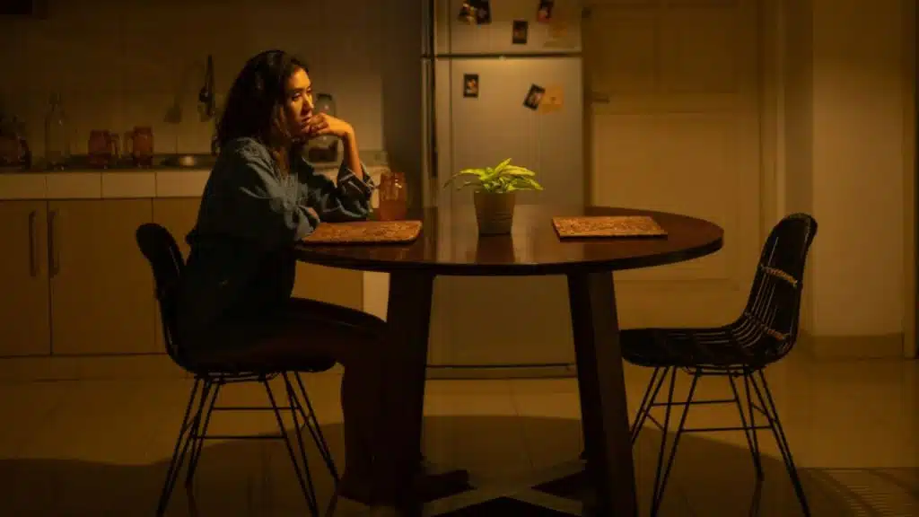 A woman sits alone in a dimly lit kitchen at a round table, resting her head on her hand.