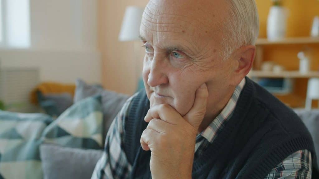 Elderly man in a plaid shirt and vest sitting with his hand on his chin.