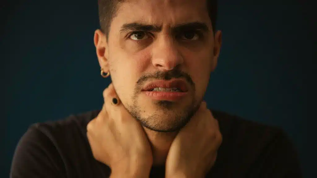 Man with beard and dark shirt clutches his neck, making a distressed face.