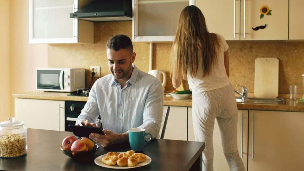 A man in a light blue shirt smiles while using a tablet at a kitchen table as a woman faces away, working in the background.