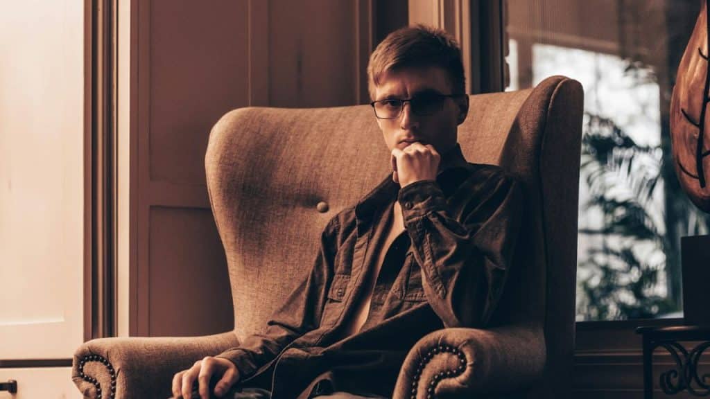 Young man in glasses sitting in a large armchair, chin resting on his hand.