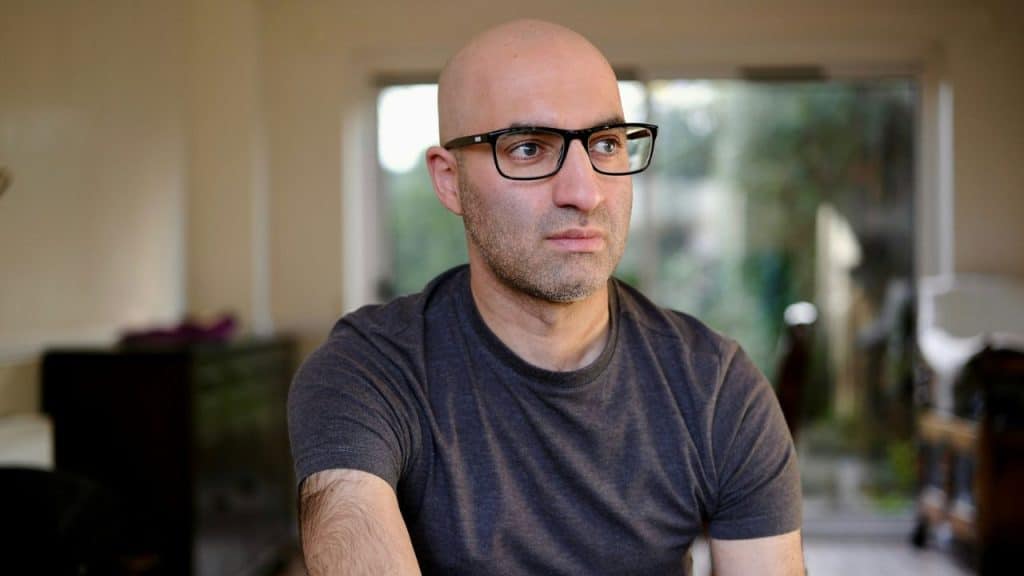 A bald man with glasses and a gray shirt looks off-camera indoors.
